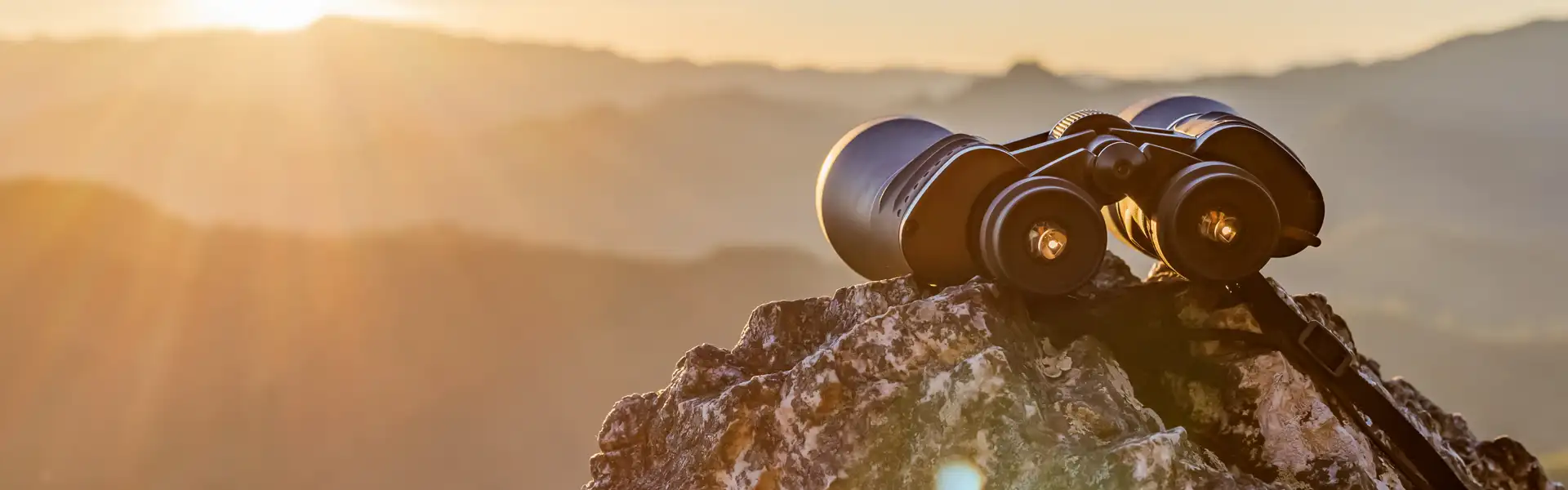 A pair of binoculars rests on a rock, with a mountain landscape in the background and sunbeams shining through the air